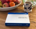 box of Covid-19 rapid tests on a coffee table in front of a bowl of fruit and a hot drink