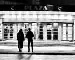 A black and white photo taken from behind two people standing looking into the Plaza cinema entrance on opening night December 2023