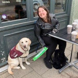 lady sat outside a café with her guide dog. She's smiling at the camera