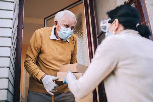 lady wearing a facemask passing a boxed meal to an elderly man at his front door